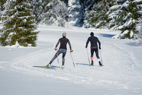Zwei Skilangläufer auf einer schneebedeckten Strecke in einer winterlichen Landschaft.